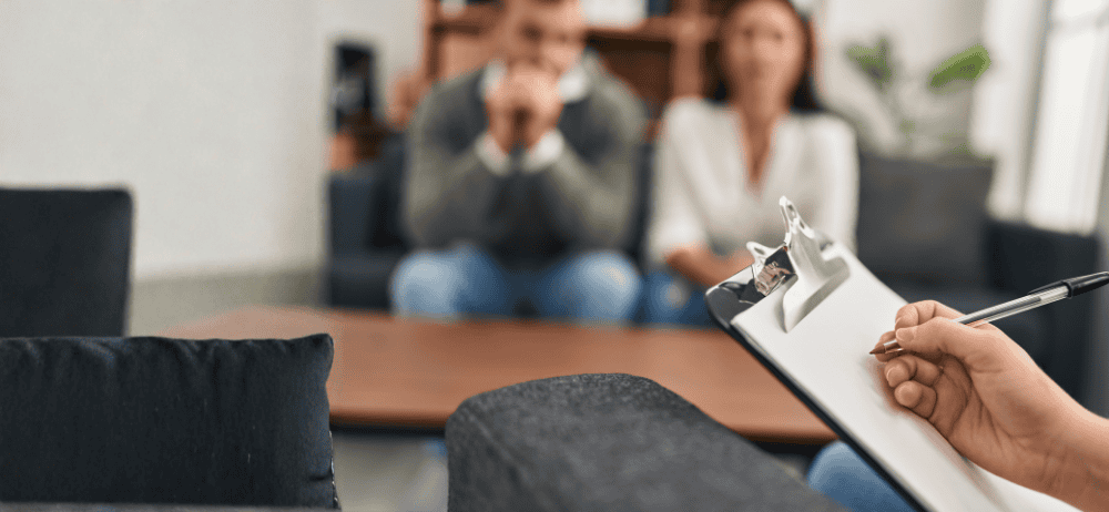 Therapist holding clipboard with blurred couple on a couch