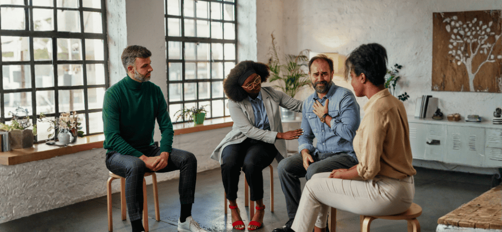 Ethical non monogamy partners in a therapy session. One partner is overwhelmed with emotion while another comforts him. Tow others sit and watch. 