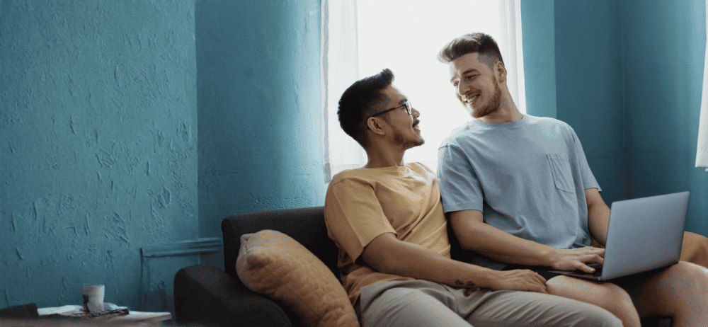 Two men sitting on a couch together with a laptop while they smile at each other